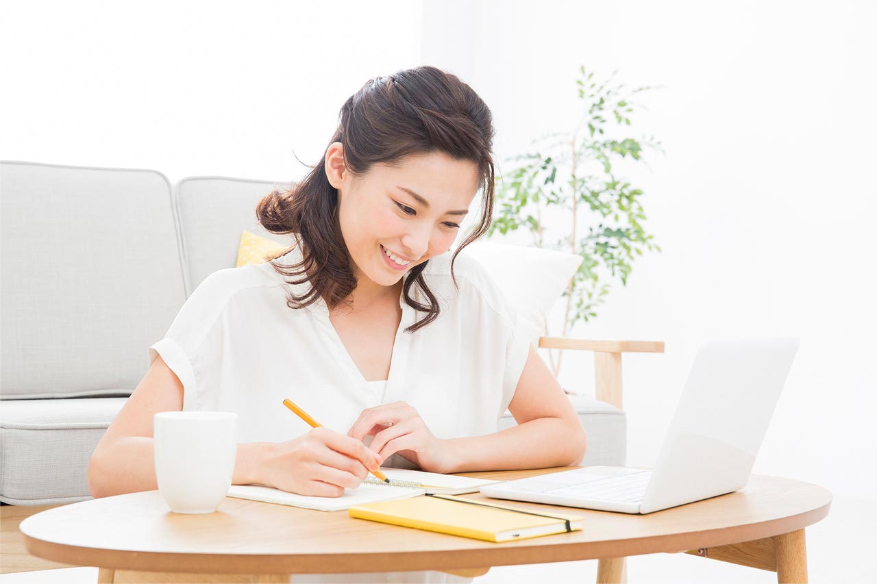 ノートパソコンを開いてペンで紙に何か書いている女性